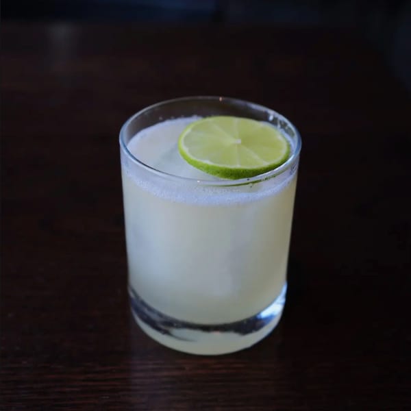 A light-colored cocktail with a lime slice garnish sits on a dark wooden table.