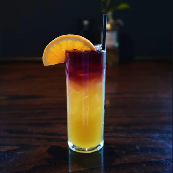 A vibrant layered cocktail with orange and red hues, garnished with an orange slice and black straw.