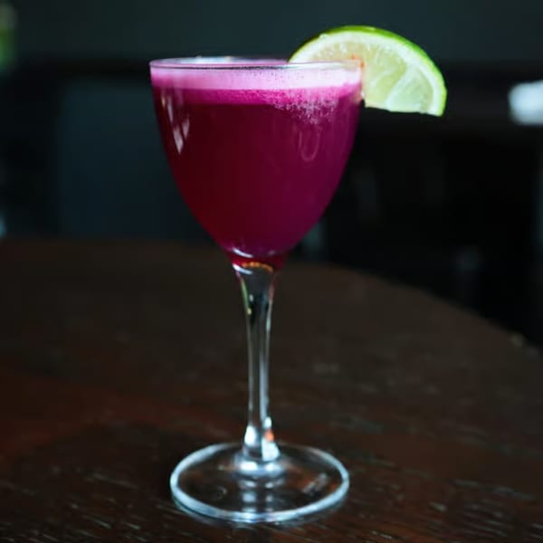 A vibrant purple cocktail with a foamy top and a lime wedge garnish, on a dark wooden table.
