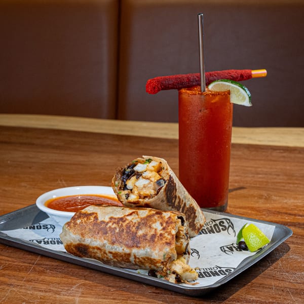 Two Sonora burritos on a tray with a side of salsa, lime wedges, and a Bloody Mary.