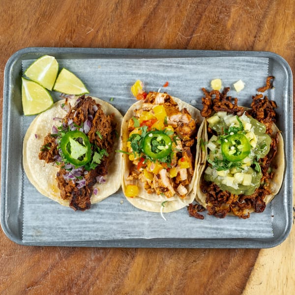 Three Sonora Mexican Kitchen tacos, including shredded beef, chicken, and al pastor, with lime wedges.