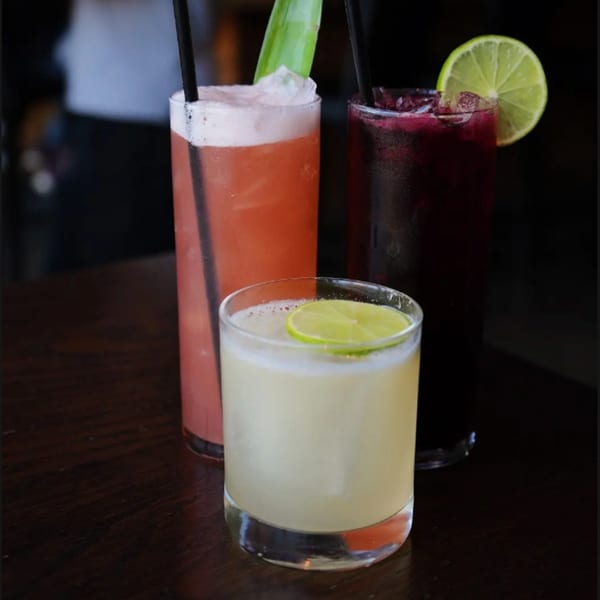 Three colorful cocktails on a dark wooden table, garnished with lime and a green leaf.