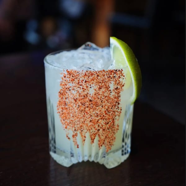 A cocktail with ice, a lime wedge, and chili salt on the rim, on a dark wooden surface.