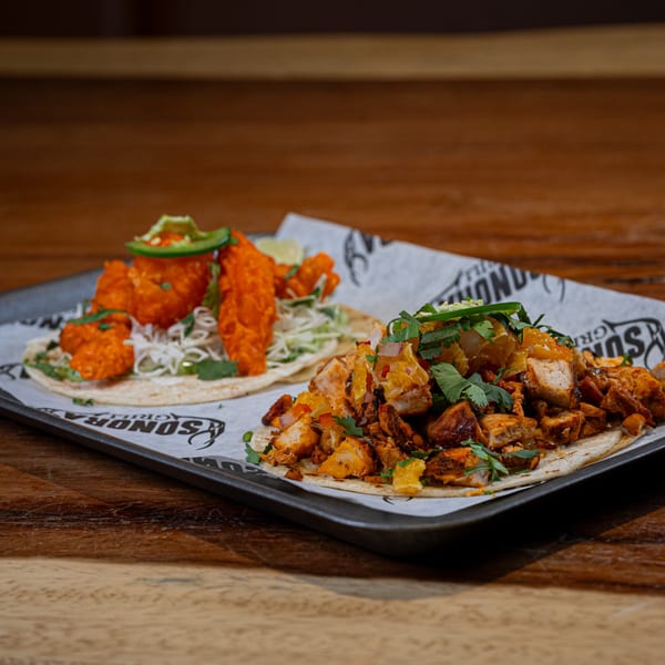 Two Sonora Mexican Kitchen tacos on a tray, one with orange chicken, the other with diced meat.