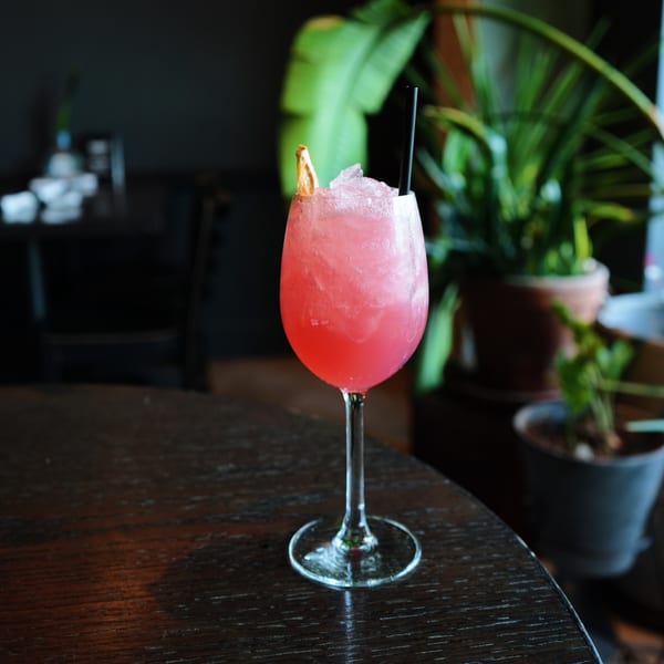 Pink cocktail with ice, a black straw, and an orange slice on a dark wooden table.