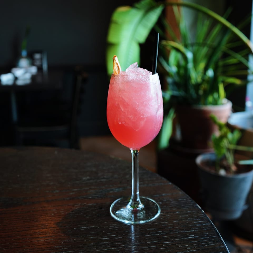 Pink cocktail with ice, a black straw, and an orange slice on a dark wooden table.