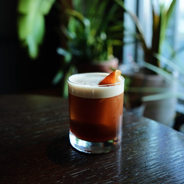 A Sonora Mexican Kitchen cocktail with foamy top and orange slice, on a dark wooden table.