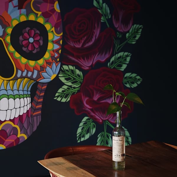 A vibrant sugar skull mural and red roses adorn a dark wall, with a plant in a bottle on a wooden table.