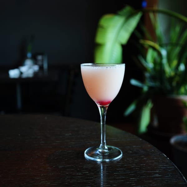 Pink cocktail with a red cherry at the bottom, on a dark wooden table.