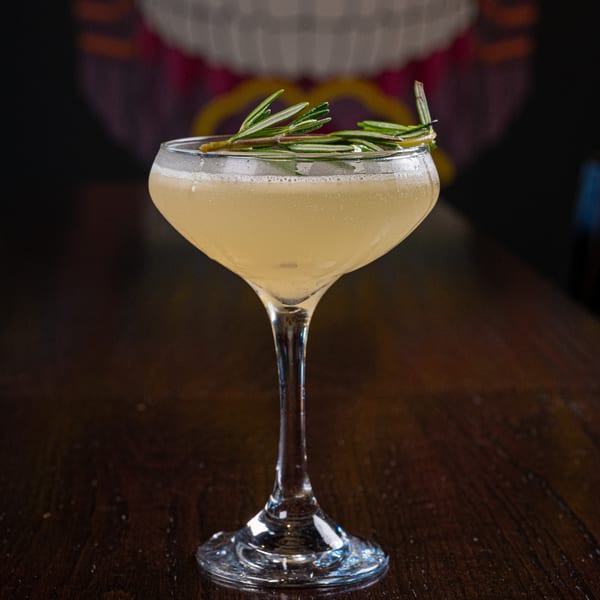 A light yellow cocktail in a coupe glass, garnished with fresh rosemary sprigs on a dark wooden table.